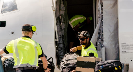 Airport ground crew members in high-visibility gear loading luggage into an airplane cargo hold.