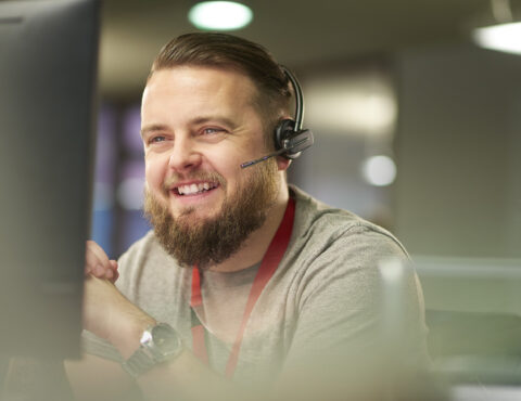 A man with a beard and headset sits at a desk in a modern office. He smiles while looking at a computer screen, wearing a gray shirt with a red lanyard.