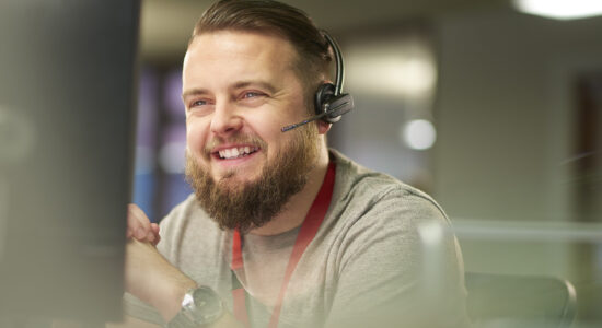 A man with a beard and headset sits at a desk in a modern office. He smiles while looking at a computer screen, wearing a gray shirt with a red lanyard.