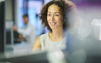A woman with curly hair, wearing a headset and a light gray top, is engaged in a phone conversation. She is smiling while sitting in an office environment with blurred background details.