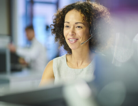A woman with curly hair, wearing a headset and a light gray top, is engaged in a phone conversation. She is smiling while sitting in an office environment with blurred background details.