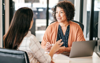 Two women in a modern office setting have a conversation. One woman, wearing an orange cardigan, is holding a pen and speaking, while the other woman, with long dark hair, listens attentively.