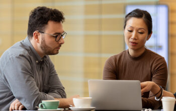 Three professionals—two women and one man—sit at a table in a modern office environment. The woman on the right is pointing to a laptop screen while explaining something to her colleagues.
