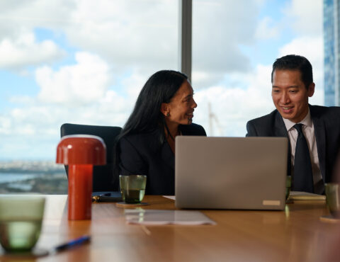 Two people sitting down in a corporate office. In the background in a blurred view of a city and blue cloudy sky.