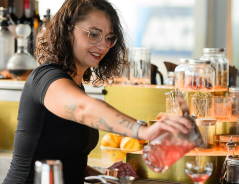 A smiling bartender with curly hair, glasses, and tattoos on her arms is pouring a red drink into a glass. The bar is well-stocked with bottles, jars, and citrus fruits in the background.