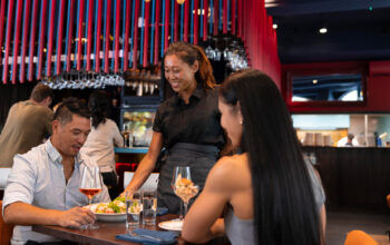 A restaurant server smiles while delivering food to a seated couple. The woman is wearing a sleeveless grey top, and the man has short hair and is holding a glass of rosé wine. The restaurant has a modern, red-accented design with hanging wine glasses.