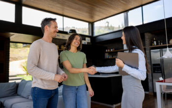 A smiling couple is shaking hands with a real estate agent holding a laptop. They stand inside a bright, modern house with large windows and wooden finishes.