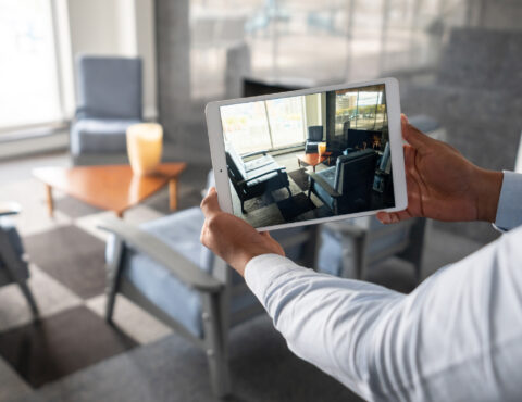 A person holds a tablet displaying a furnished living room. The tablet screen mirrors the room's layout, which features armchairs, a coffee table, and large windows.