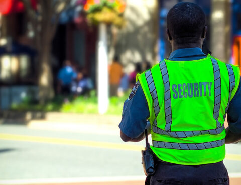 A security guard in a neon green vest with reflective stripes stands outdoors during the day, facing away from the camera. The word 