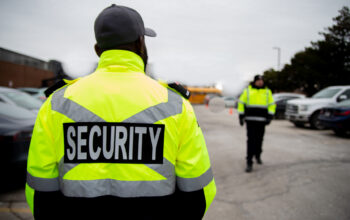 A security guard in a high-visibility neon yellow jacket stands with their back to the camera. The jacket has 