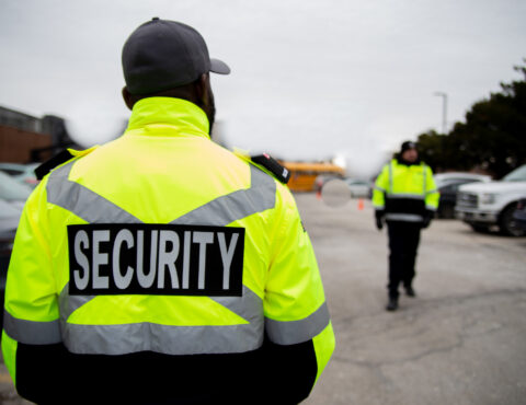 A security guard in a high-visibility neon yellow jacket stands with their back to the camera. The jacket has 