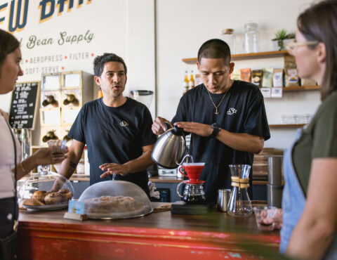 Four people in a coffee shop. One is pouring hot water in to a glass jug.