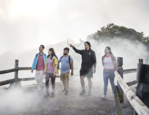 A group of five people walk along a foggy geothermal path bordered by wooden fences. One person, wearing a black jacket, points ahead while leading the group. The others wear casual outdoor clothing and backpacks, appearing engaged as they look in the same direction.