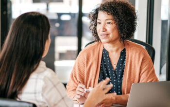 Two women engaged in conversation at a table in a professional setting. The woman on the right, wearing an orange cardigan, smiles attentively while holding a pen. The other woman, seen from behind, has long dark hair and is gesturing with her hand. A laptop is visible on the table.