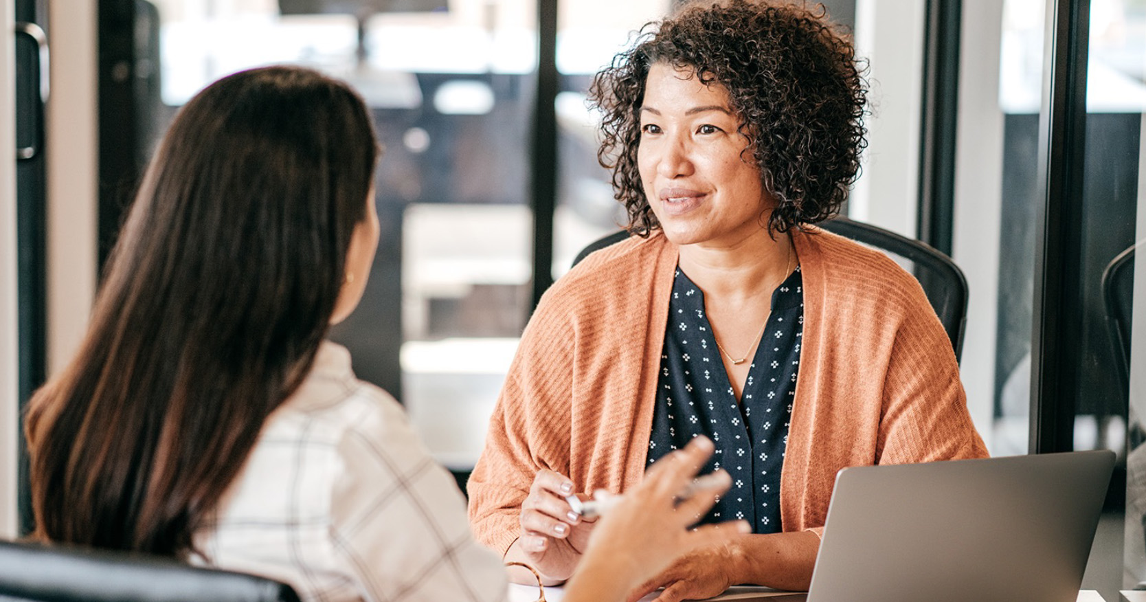 Two women engaged in conversation at a table in a professional setting. The woman on the right, wearing an orange cardigan, smiles attentively while holding a pen. The other woman, seen from behind, has long dark hair and is gesturing with her hand. A laptop is visible on the table.