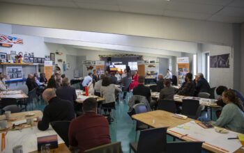 A wide view of a workshop or meeting with participants seated at tables, facing a group of speakers at the front. Ringa Hora banners and posters are displayed around the room.