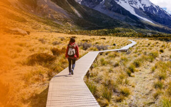 A person walking on a wooden path leading towards the mountains. They are wearing a red jacket and backpack. On the right is an orange gradient.