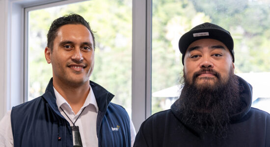A close up of two people looking towards the camera. One is wearing a blue vest and white shirt. The other is wearing a cap and jumper.