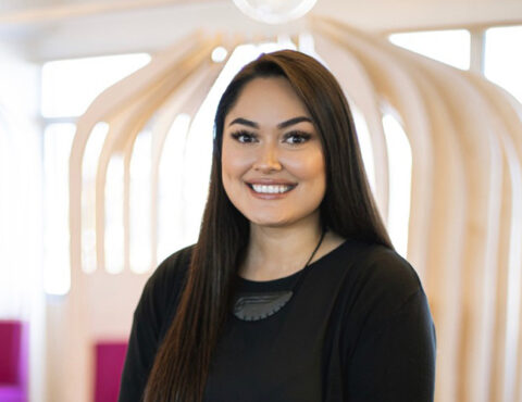 A woman with long dark hair smiles warmly while standing in a brightly lit room with modern wooden architectural features in the background. She is wearing a black top and a carved necklace, exuding a professional yet approachable demeanor.