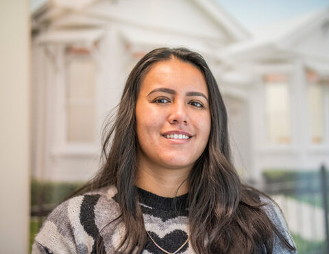 A close-up portrait of a smiling individual with long dark hair wearing a patterned sweater. The background features a softly blurred image of a traditional white house, creating a warm and inviting atmosphere.