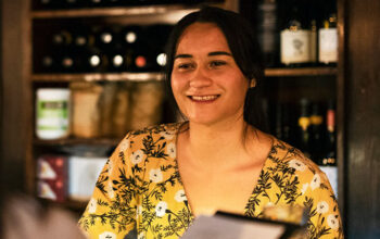A smiling woman wearing a yellow floral-patterned dress standing behind a counter, with shelves of wine bottles and other items in the background, creating a warm and welcoming atmosphere.