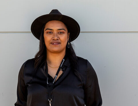A portrait of a confident individual standing against a neutral light grey background. They are dressed in a sleek black shirt and a wide-brimmed black hat, accessorized with subtle gold jewelry and a lanyard. The image captures a poised and professional demeanor.