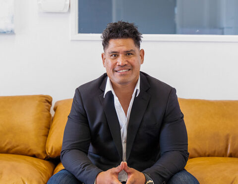 A man wearing a dark blazer and white shirt sits on a tan couch in a modern, minimalistic setting. He is smiling confidently and clasping his hands, with a light-colored wall and window in the background.