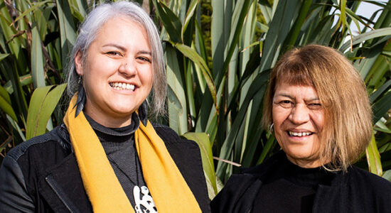 Two wahine Māori (Māori women) standing in front of a flax bush.