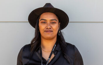 A portrait of a confident individual standing against a neutral light grey background. They are dressed in a sleek black shirt and a wide-brimmed black hat, accessorized with subtle gold jewelry and a lanyard. The image captures a poised and professional demeanor.