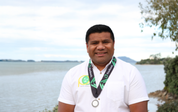 A man wearing a white t-shirt, and a medal around his neck from Kiwibank. In the background is an ocean.