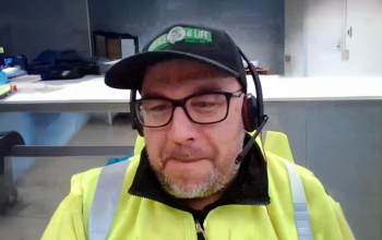 A man wearing a bight yellow jacket and cap.He is sitting in an office and looking at a compute screen.
